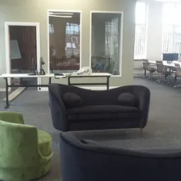 Wnętrze biura z zielonym krzesłem i ciemnoszarą kanapą na pierwszym planie, biurka z komputerami i krzesłami w tle.