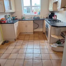 Ustawna kuchnia z beżowymi frontami szafek i brązową terakotą na podłodze, widok na okno i sprzęty AGD, mop oparty o ścianę.