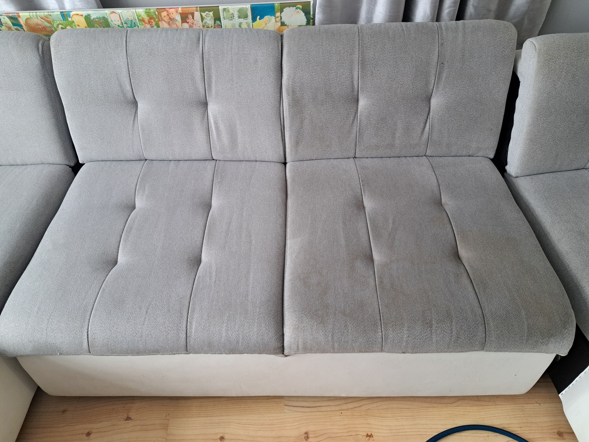 Szara, tapicerowana kanapa z widocznymi śladami użytkowania na siedziskach, ukazana w dziennym świetle, z fragmentem podłogi i zasłon w tle.