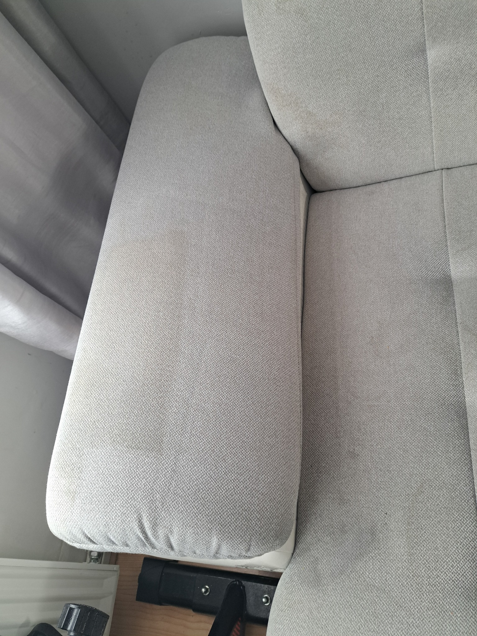 Fragment jasnoszarej, tekstylnej sofy z widocznymi plamami na podłokietniku przed czyszczeniem, z fragmentem zasłony w tle.