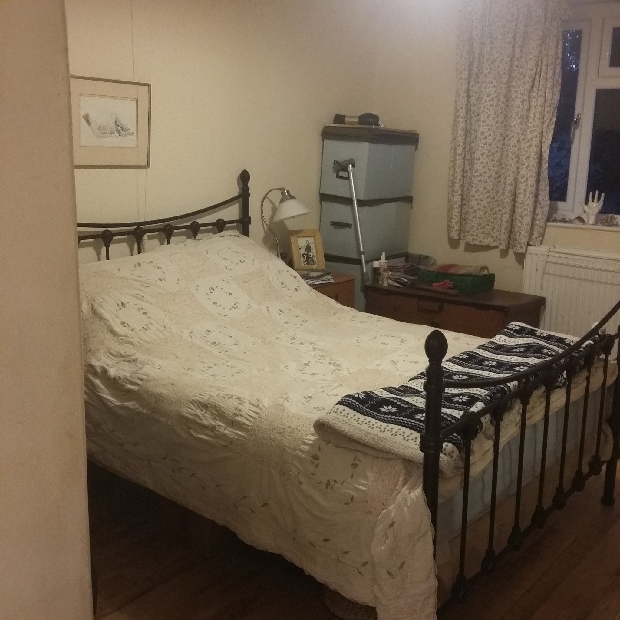 Pokój z łóżkiem z metalową ramą, nakrytym kremową narzutą z haftem i wzorzystym kocem, obok szafka nocna z lampką i zdjęciem w ramce, w tle okno z zasłoną.