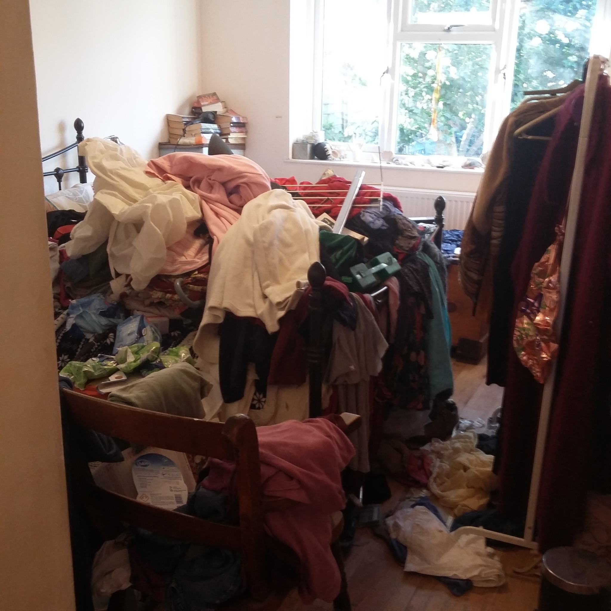 Pokój zagracony ubraniami i różnymi przedmiotami, z widocznym łóżkiem, krzesłem i suszarką na pranie.