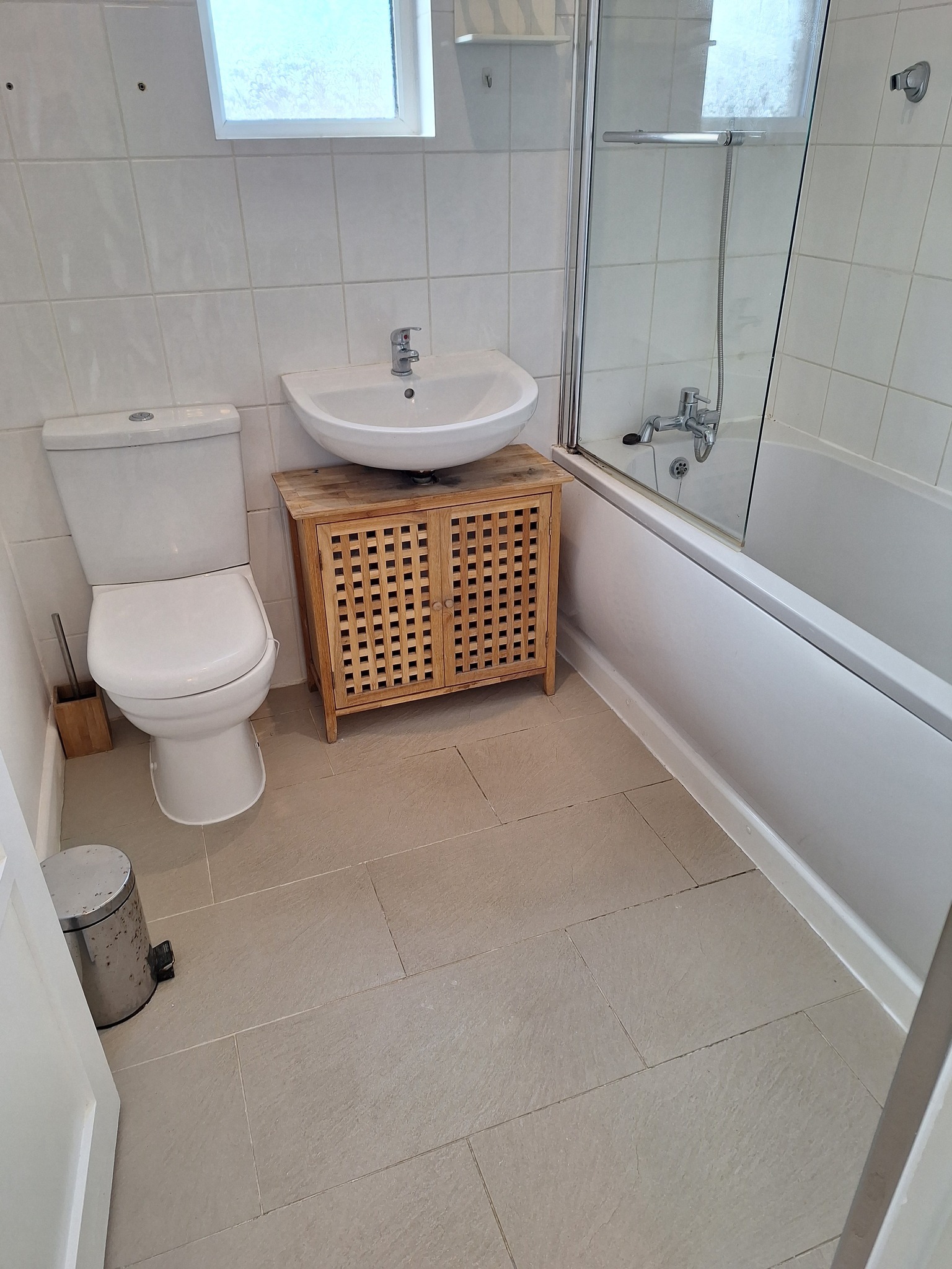 Umywalka z szafką z drewnianą kratką, toaleta i wanna z parawanem w łazience z jasnymi płytkami.