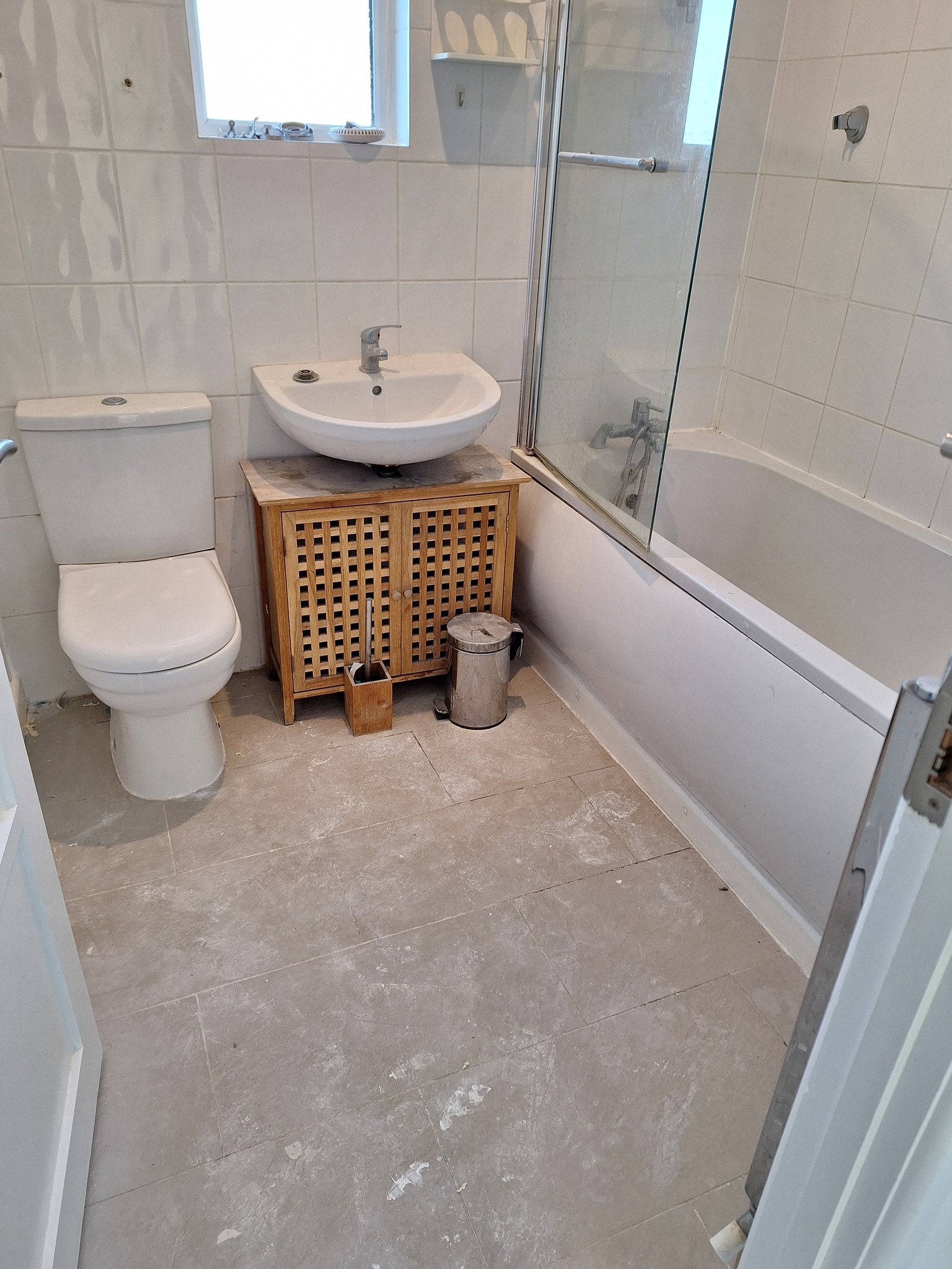 Biała łazienka z umywalką na drewnianej szafce, toaletą i wanną z parawanem, widoczne ślady po remoncie na podłodze.