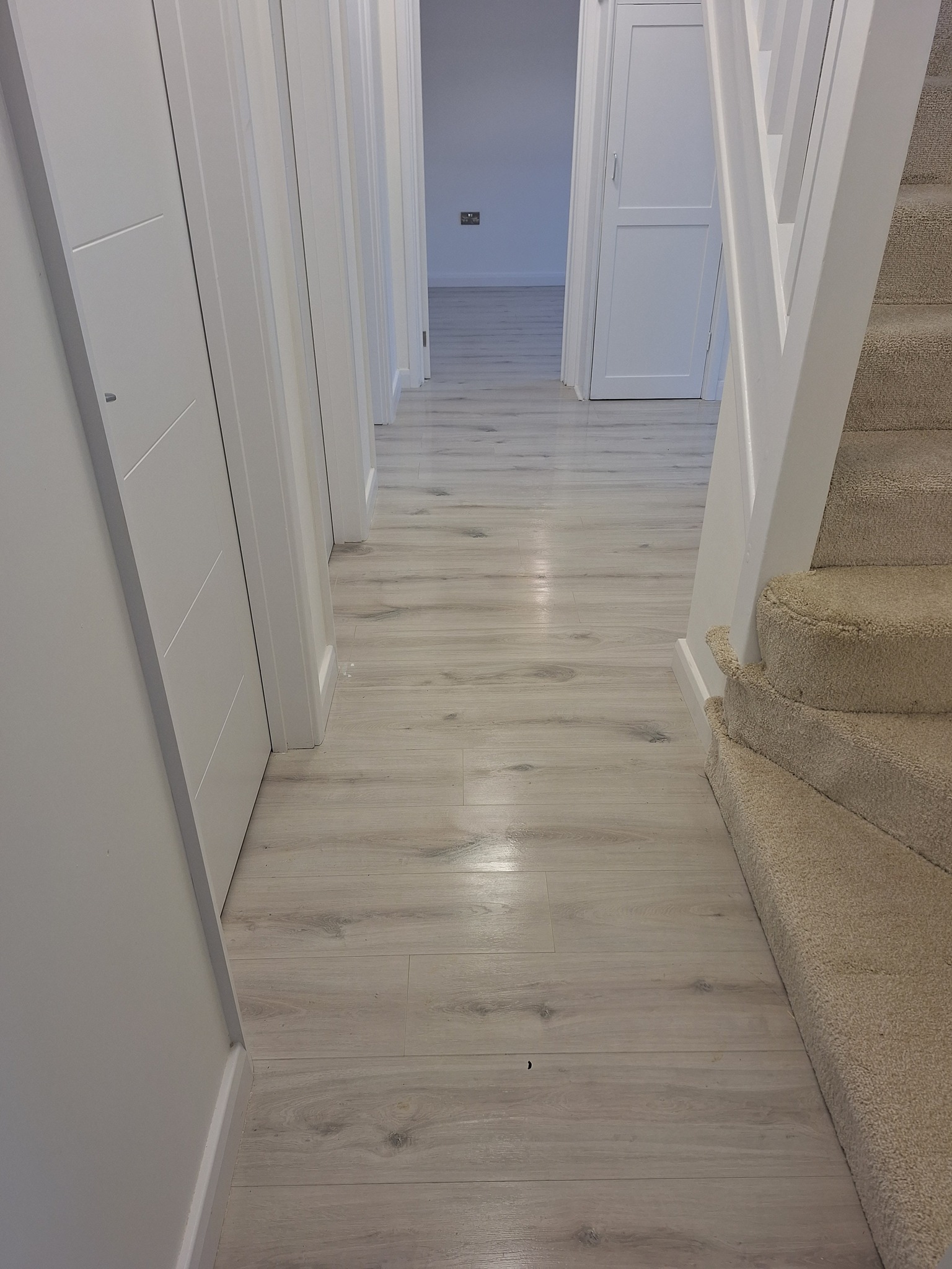 Jasne panele podłogowe w korytarzu z białymi drzwiami i schodami wyłożonymi beżową wykładziną.