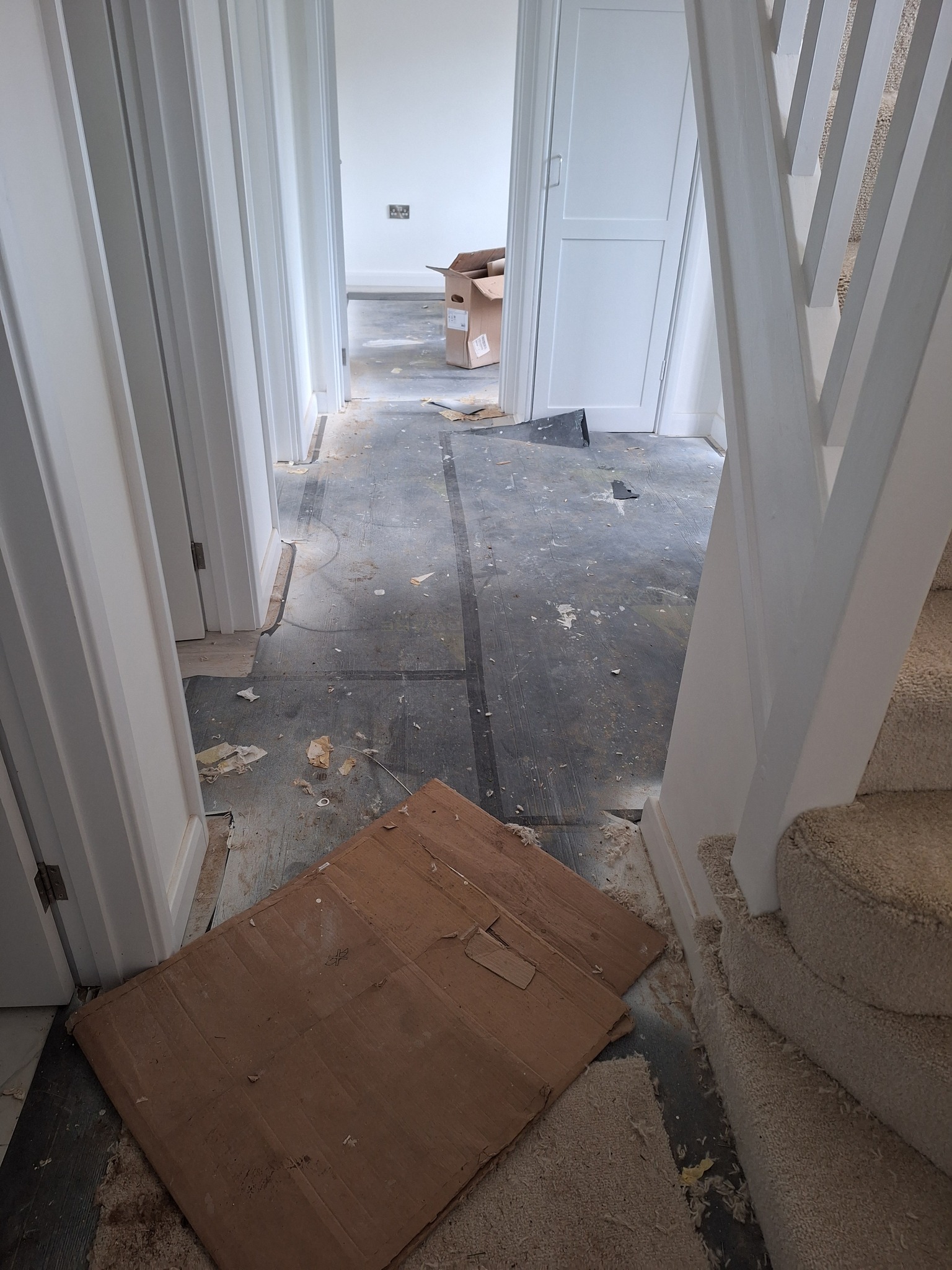 Korytarz w trakcie remontu: podłoga częściowo zabezpieczona kartonem, widoczne ślady po zrywaniu starej podłogi, białe drzwi i schody.