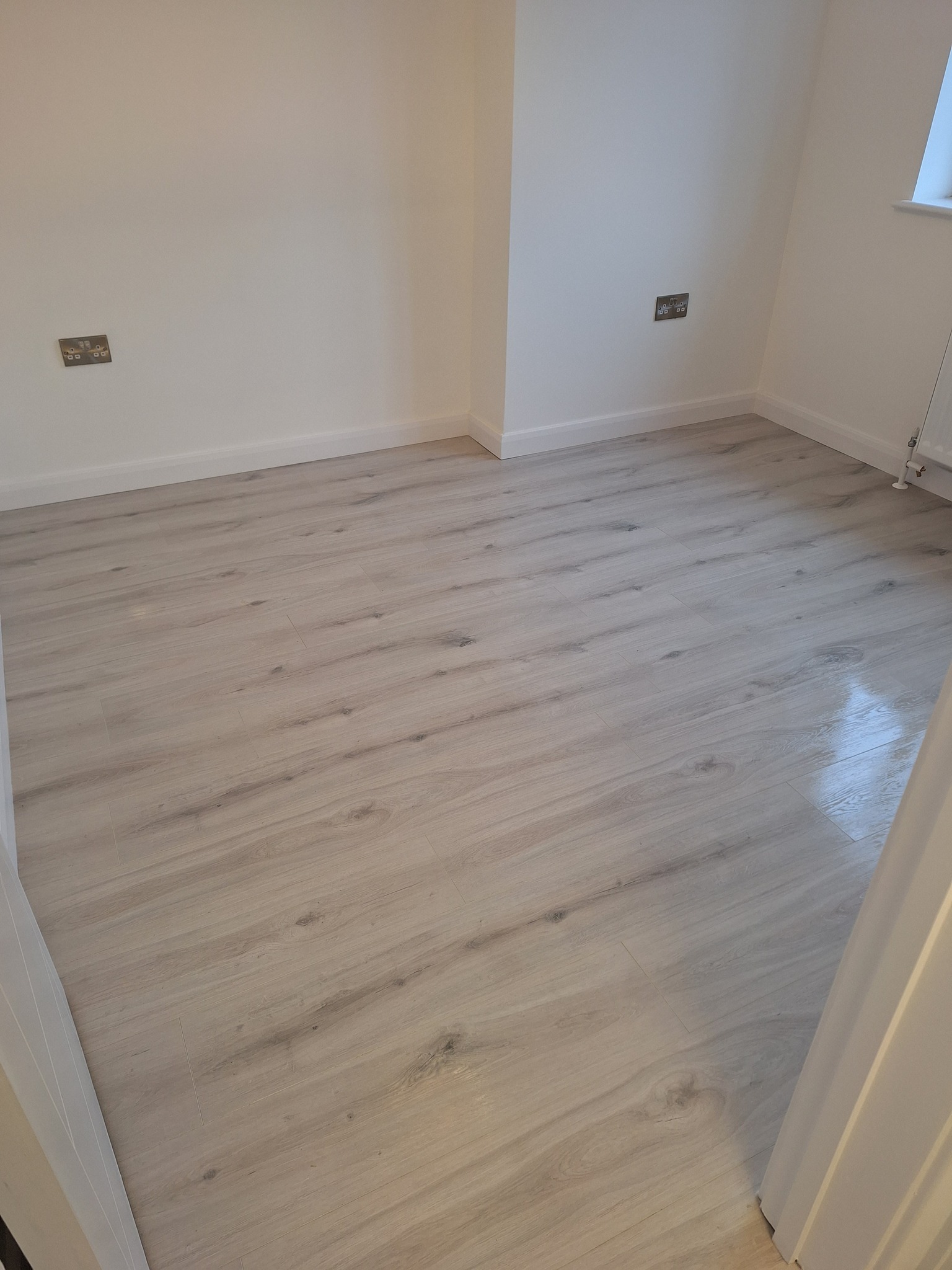 Jasna, pusta przestrzeń z panelami podłogowymi imitującymi drewno, widoczne gniazdka elektryczne na białych ścianach i biała listwa przypodłogowa.