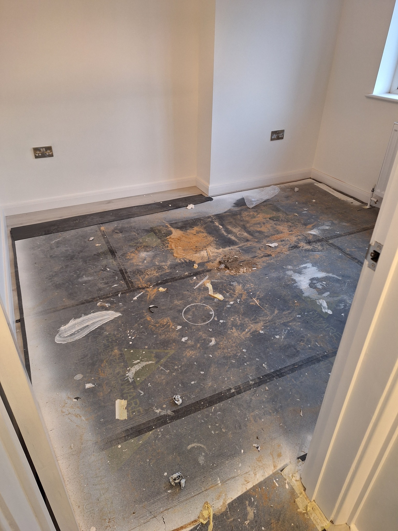 Pokój w trakcie remontu: zabezpieczona podłoga z widocznymi śladami farby, gipsu i innych zabrudzeń po pracach budowlanych, widoczne gniazdka elektryczne na ścianie.