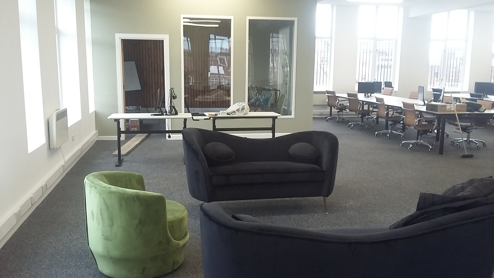 Wnętrze biura z zielonym krzesłem i ciemnoszarą kanapą na pierwszym planie, biurka z komputerami i krzesłami w tle.