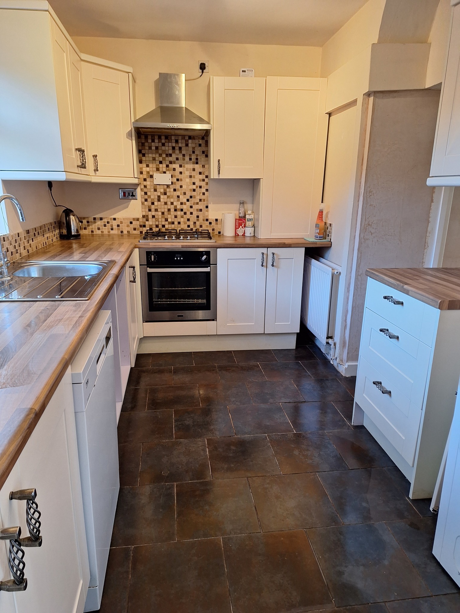 Jasna kuchnia z drewnianym blatem i mozaiką nad kuchenką, widoczne brązowe płytki podłogowe i grzejnik.