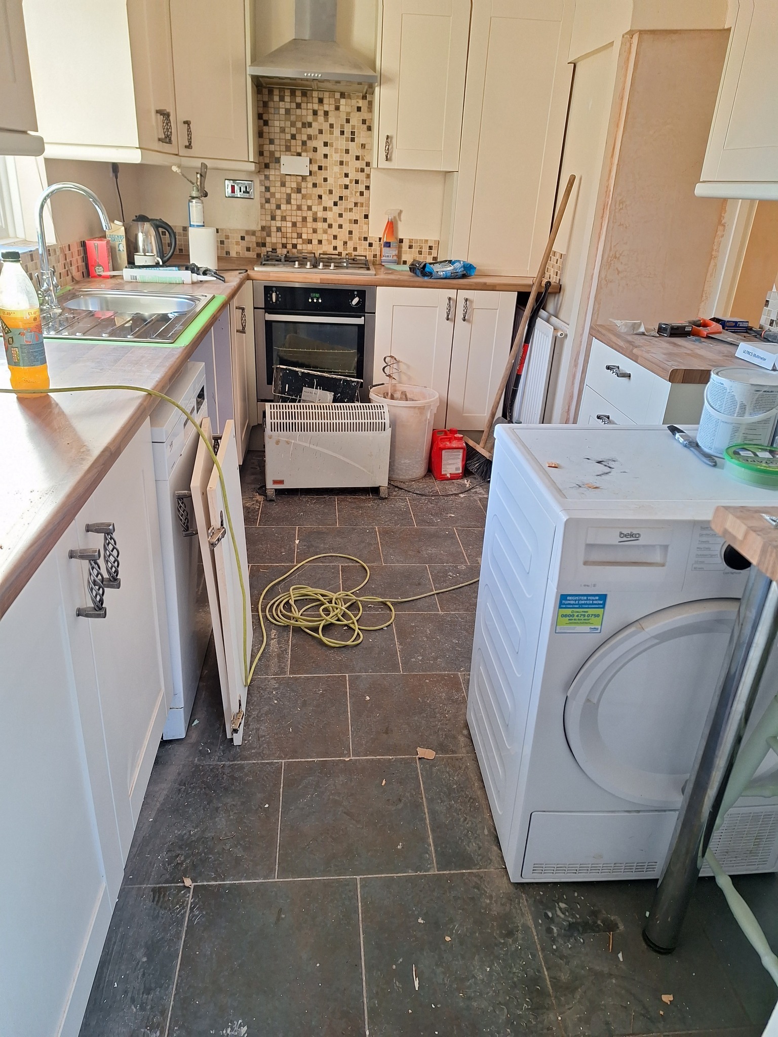 Kuchnia w trakcie remontu: widoczne niedokończone szafki, sprzęt AGD, grzejnik elektryczny, rozłożony kabel i ślady po pracach budowlanych na podłodze z płytek.