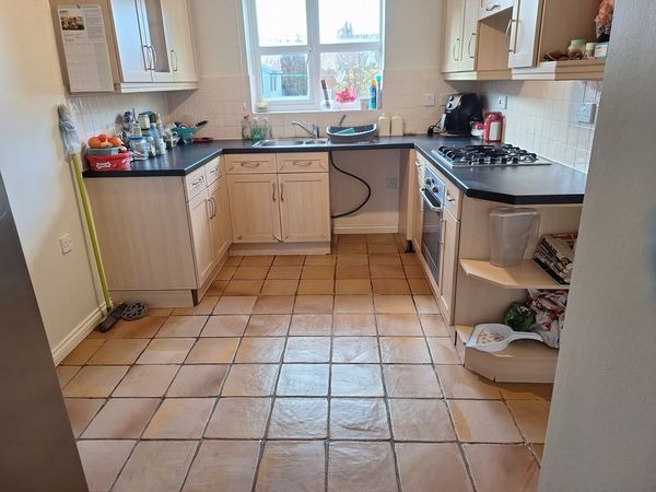 Ustawna kuchnia z beżowymi frontami szafek i brązową terakotą na podłodze, widok na okno i sprzęty AGD, mop oparty o ścianę.