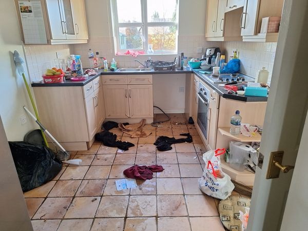 Bardzo zabrudzona kuchnia przed sprzątaniem: brudna podłoga z płytek, resztki jedzenia, porozrzucane ubrania, otwarte szafki, widok zza uchylonych drzwi.