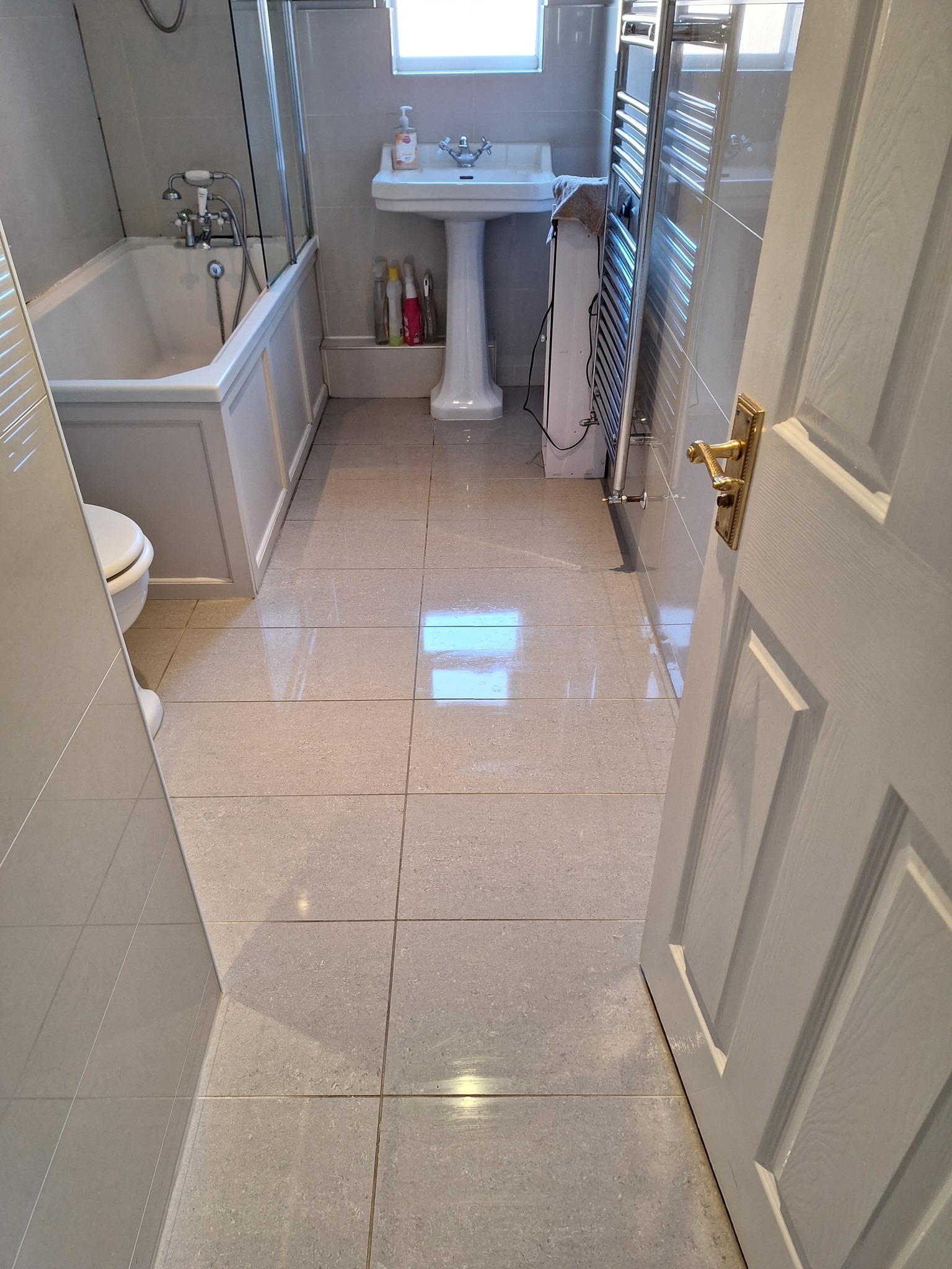 Jasna łazienka z wanną, umywalką na postumencie i grzejnikiem drabinkowym, widok zza uchylonych drzwi, odbicia światła na płytkach podłogowych.