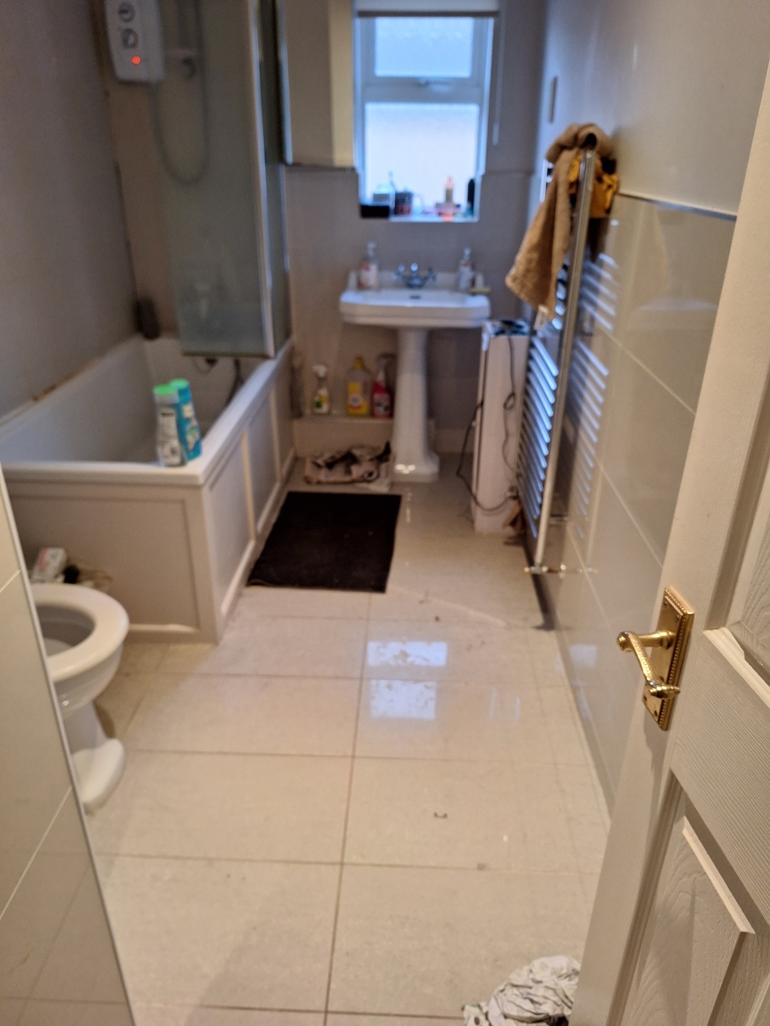 Widok na łazienkę z otwartymi drzwiami, ukazujący wannę z parawanem, sedes, umywalkę na postumencie, grzejnik drabinkowy z ręcznikiem i ciemny dywanik.