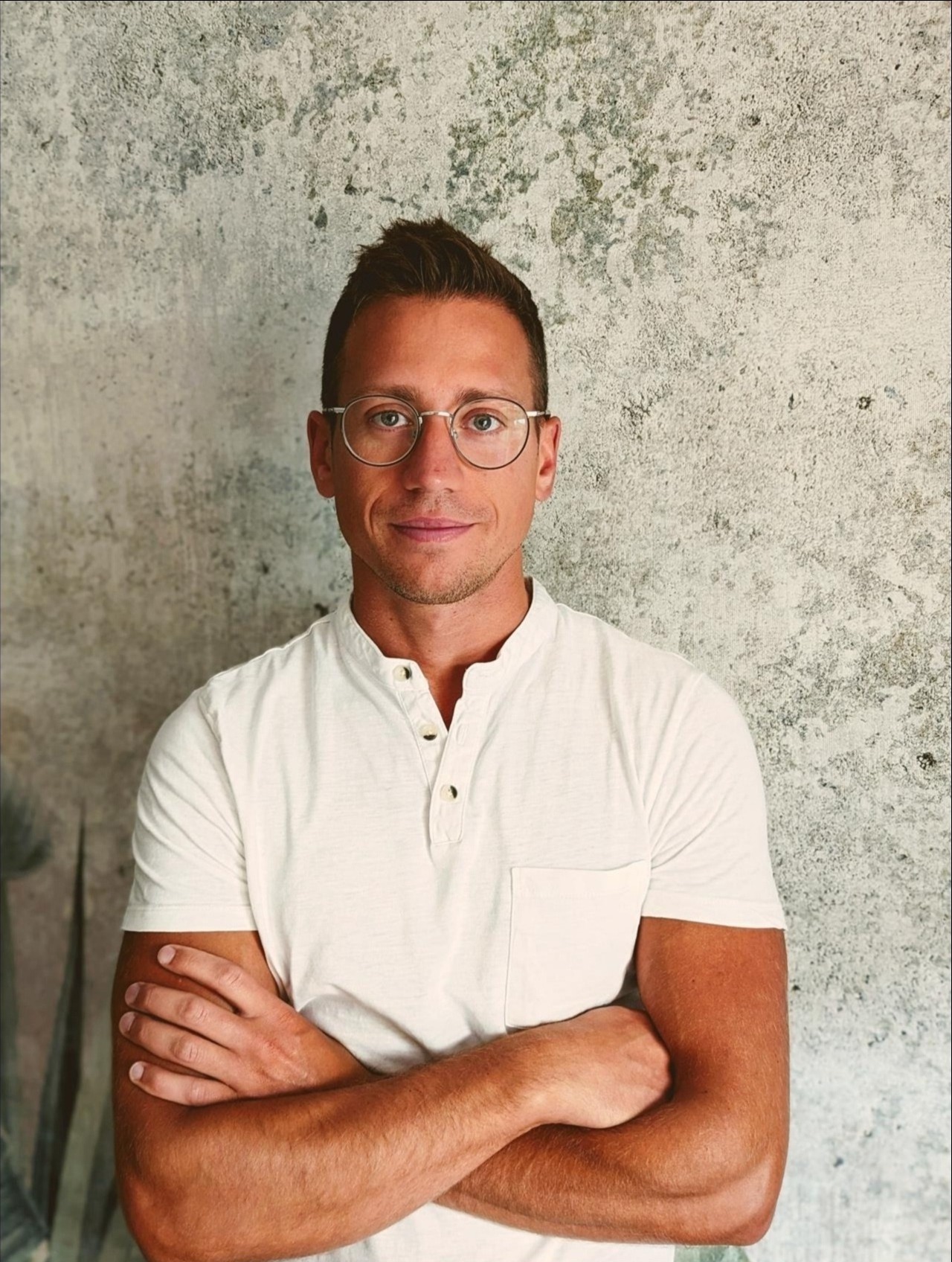 Portret mężczyzny w okularach, stojącego na tle ściany z szarą, nieregularną fakturą.