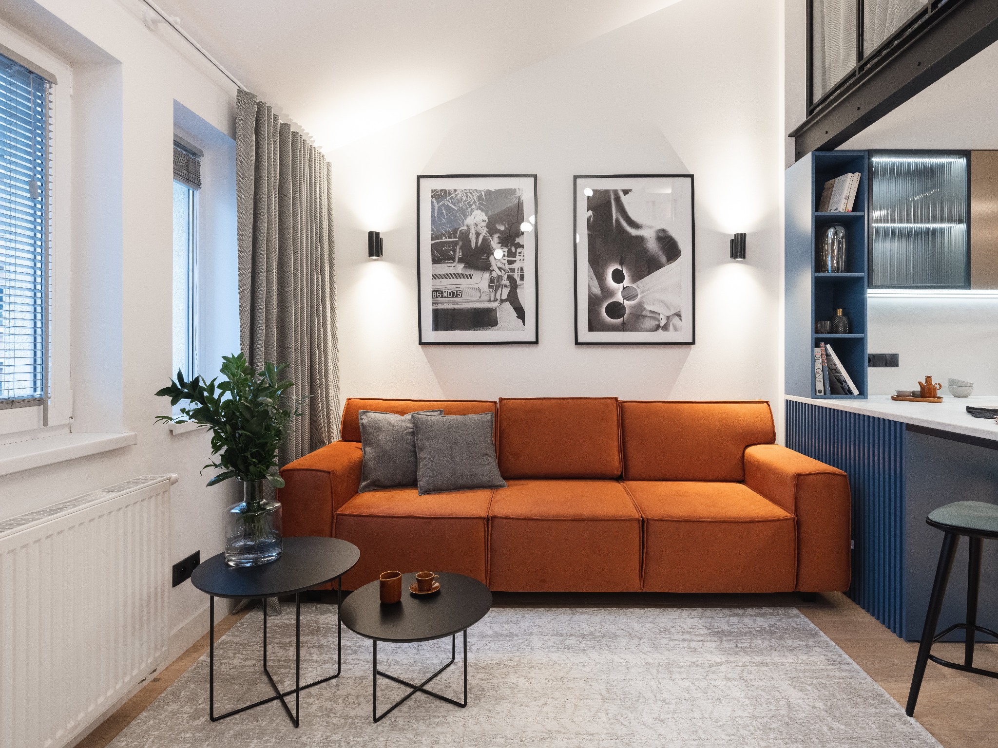 Jasne wnętrze mieszkania z pomarańczową sofą, czarnymi stolikami kawowymi i dekoracyjnymi plakatami na ścianie. Widoczne również zasłony w oknie, roślina w szklanym wazonie oraz fragment aneksu...
