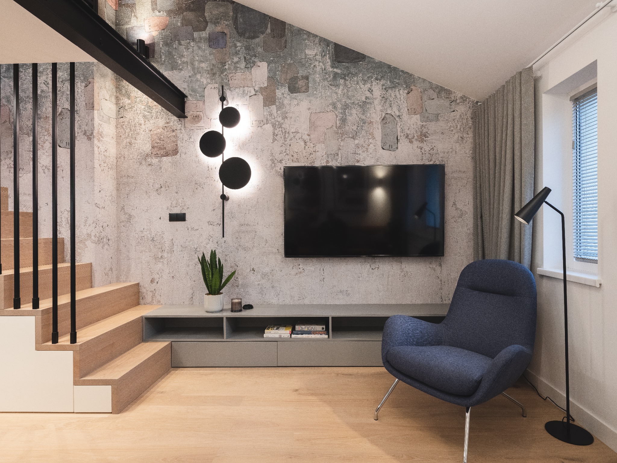 Nowoczesny salon z dekoracyjną ścianą o fakturze betonu, oświetleniem LED w kształcie kuli, drewnianymi schodami z czarnymi balustradami, szarą szafką RTV i niebieskim fotelem.