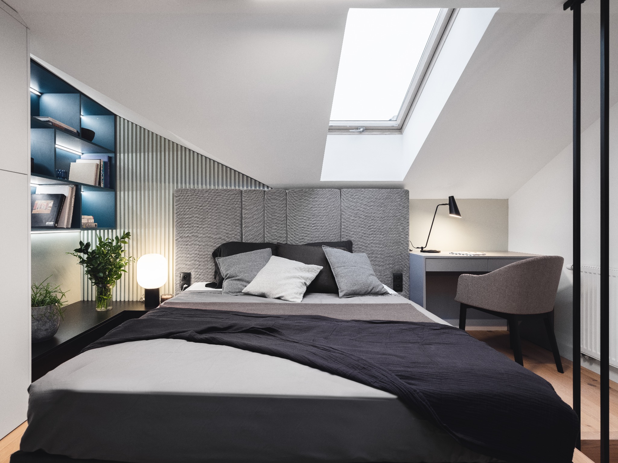 Nowoczesna sypialnia na poddaszu z łóżkiem w odcieniach szarości, biurkiem pod oknem dachowym i regałem z podświetleniem LED.