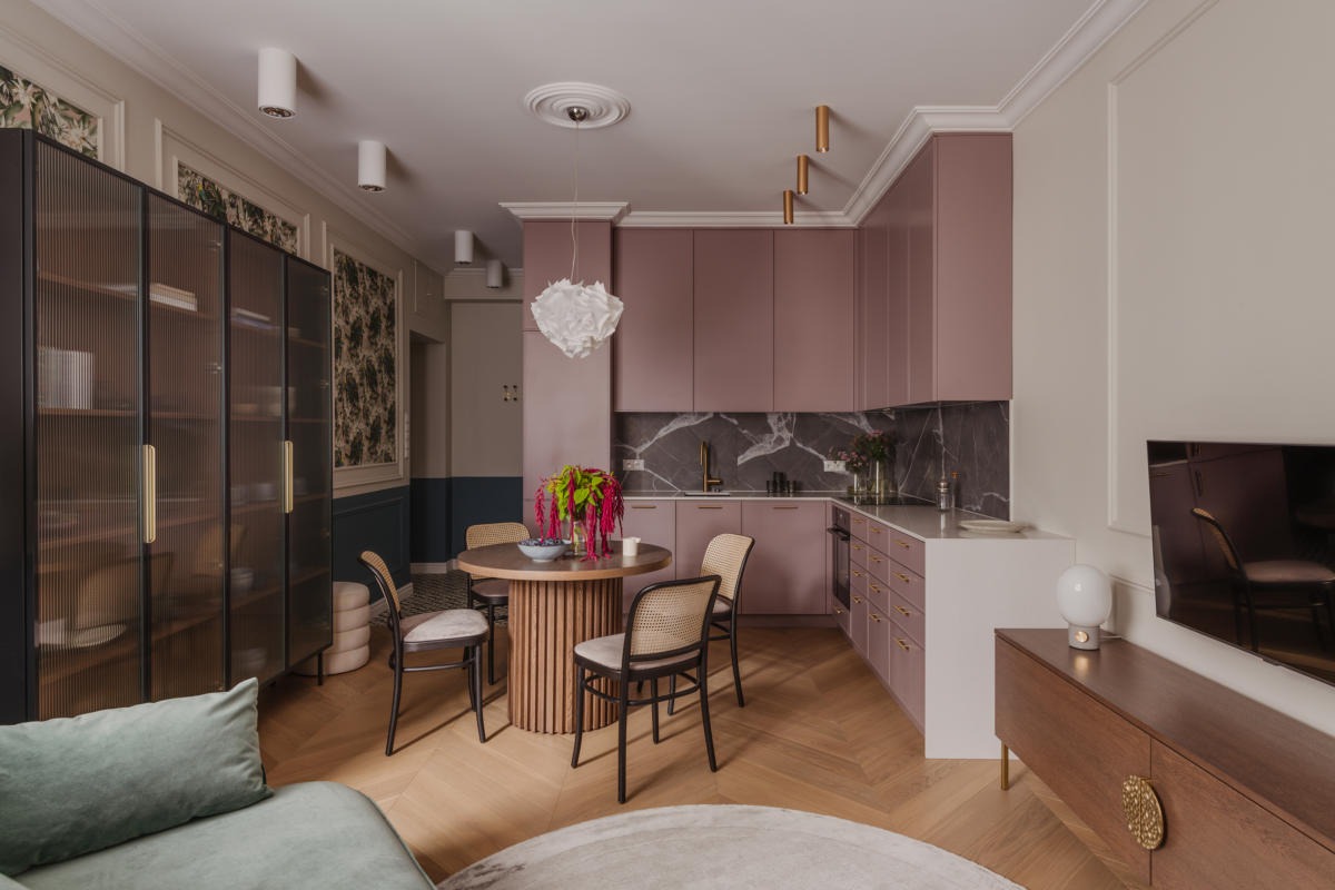 Wnętrze mieszkania z różową zabudową kuchenną, okrągłym stołem z kwiatami i parkietem ułożonym w jodełkę.