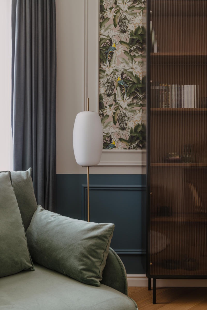 Elegancki salon z tapetą w tropikalne wzory, lampą podłogową w kształcie jajka i zieloną sofą z poduszkami, wykończony w ciemnej tonacji niebieskiego koloru.