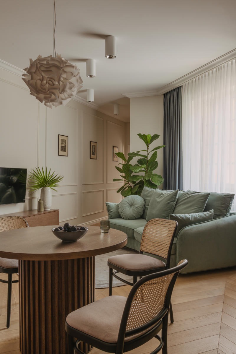 Jasne wnętrze salonu w stylu klasycznym z drewnianą podłogą w jodełkę, zieloną sofą, okrągłym stołem z ciemnymi elementami i dekoracyjnymi panelami ściennymi.