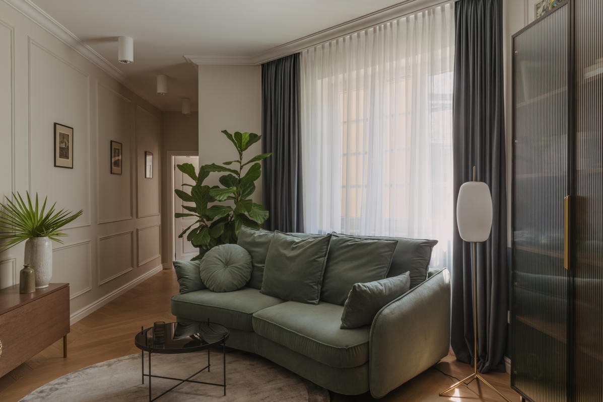 Elegancki salon z zieloną sofą, drewnianą podłogą i ścianami z dekoracyjnymi listwami, doświetlony naturalnym światłem wpadającym przez białe, zwiewne zasłony.