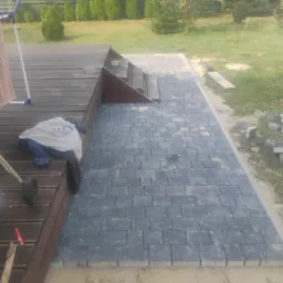 Niebiesko-szara kostka brukowa ułożona na ścieżce ogrodowej, z widocznymi niedokończonymi fragmentami i stertą kostek obok trawnika, obok tarasu z grillem i suszarką na pranie.