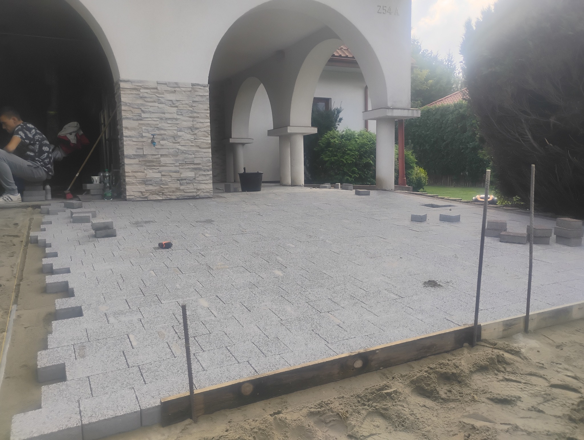 Szara kostka brukowa ułożona na podjeździe domu z łukowatymi podcieniami, widoczne niedokończone krawędzie i narzędzia pracy.