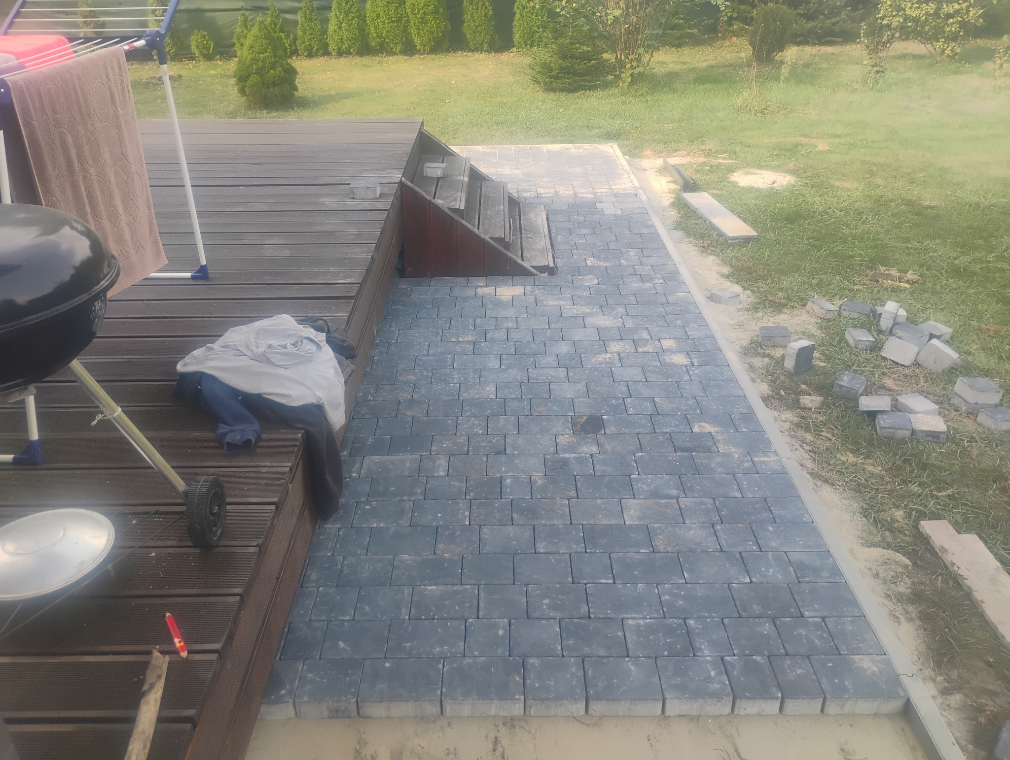Niebiesko-szara kostka brukowa ułożona na ścieżce ogrodowej, z widocznymi niedokończonymi fragmentami i stertą kostek obok trawnika, obok tarasu z grillem i suszarką na pranie.