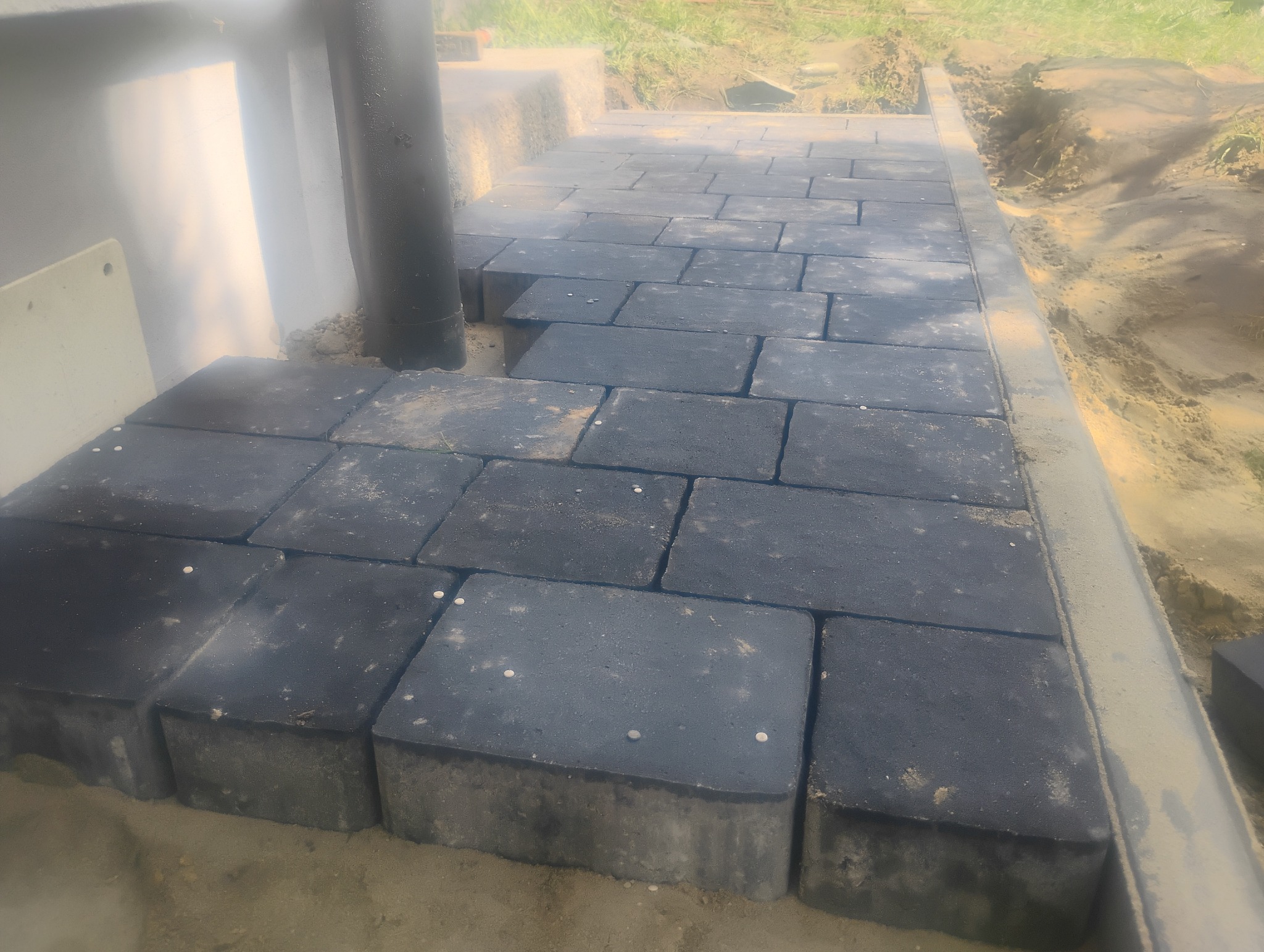Układanie szarej kostki brukowej na zewnątrz budynku, widoczne niedokończone fragmenty i rura spustowa