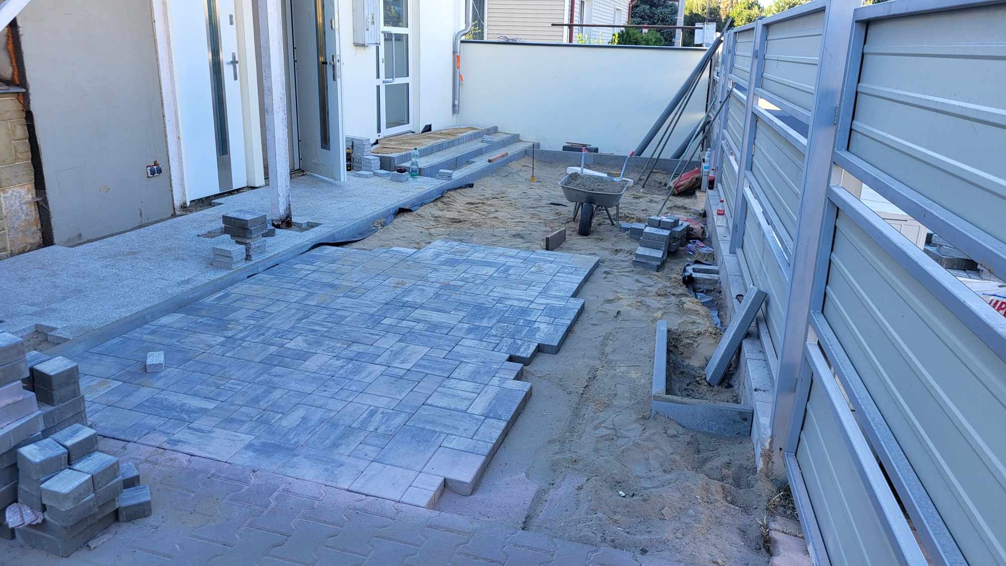 Układanie szarej kostki brukowej na podsypce piaskowej, widoczne fragmenty ułożonej nawierzchni, stos kostki, taczka z piaskiem i elementy ogrodzenia.