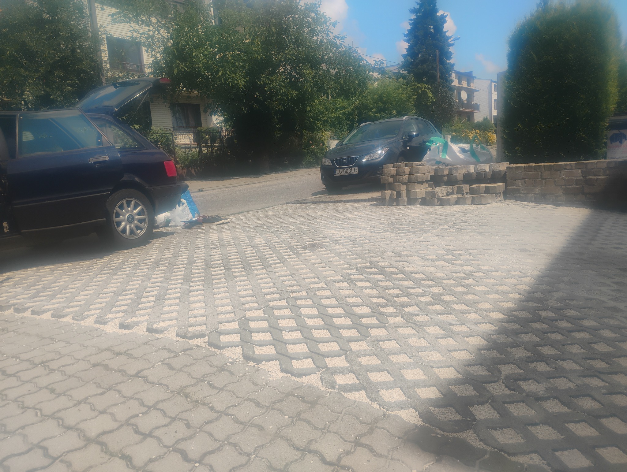 Ułożona nawierzchnia z ażurowych płyt betonowych wypełnionych kruszywem, z zaparkowanymi samochodami w tle i stertą kostki brukowej obok jednego z nich.