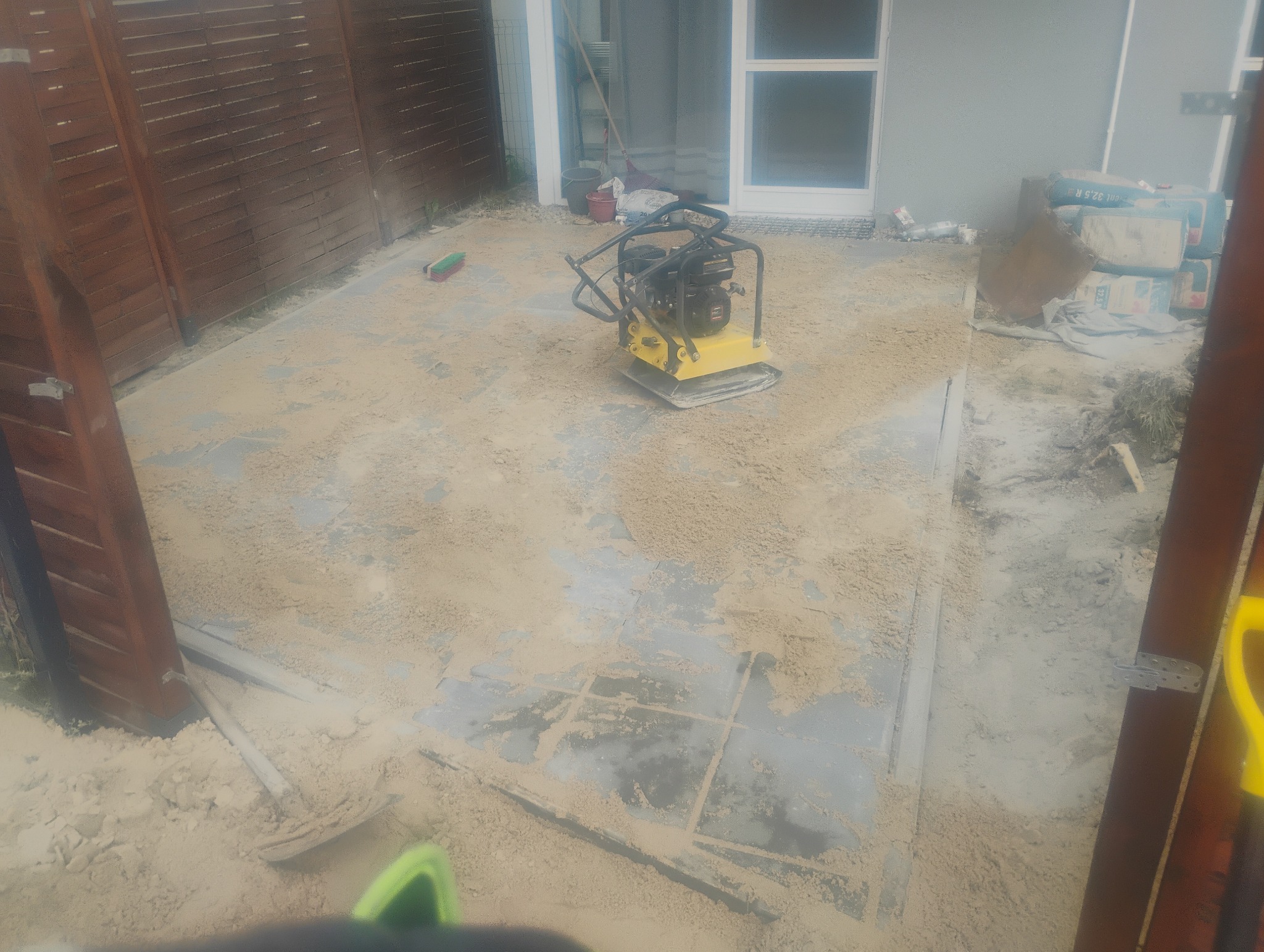 Żółta zagęszczarka na piasku, przygotowanie podłoża pod kostkę brukową na posesji z drewnianym ogrodzeniem.