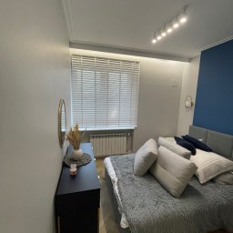 Bez Brudu 24 Dmytro Tymoshenko - Nowoczesna sypialnia z łóżkiem z szarą narzutą, ciemnoniebieską ścianą akcentującą, czarną komodą z dekoracjami i białymi żaluzjami w oknie.