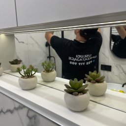 Bez Brudu 24 Dmytro Tymoshenko - Odbicie w lustrze łazienki: mężczyzna w czarnej koszulce z napisem, myjący lustro nad umywalką, na półce przed lustrem ustawione dekoracyjne sukulenty w białych doniczkach.