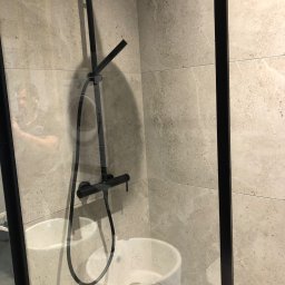 Bez Brudu 24 Dmytro Tymoshenko - Nowoczesna kabina prysznicowa z czarną armaturą na tle płytek imitujących kamień, widoczna umywalka ceramiczna za szybą.