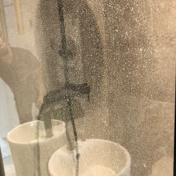 Bez Brudu 24 Dmytro Tymoshenko - Zabrudzona kabina prysznicowa z widocznymi kroplami wody i osadem, białe umywalki w tle, osoba czyszcząca widoczna jako rozmazana sylwetka po lewej stronie.