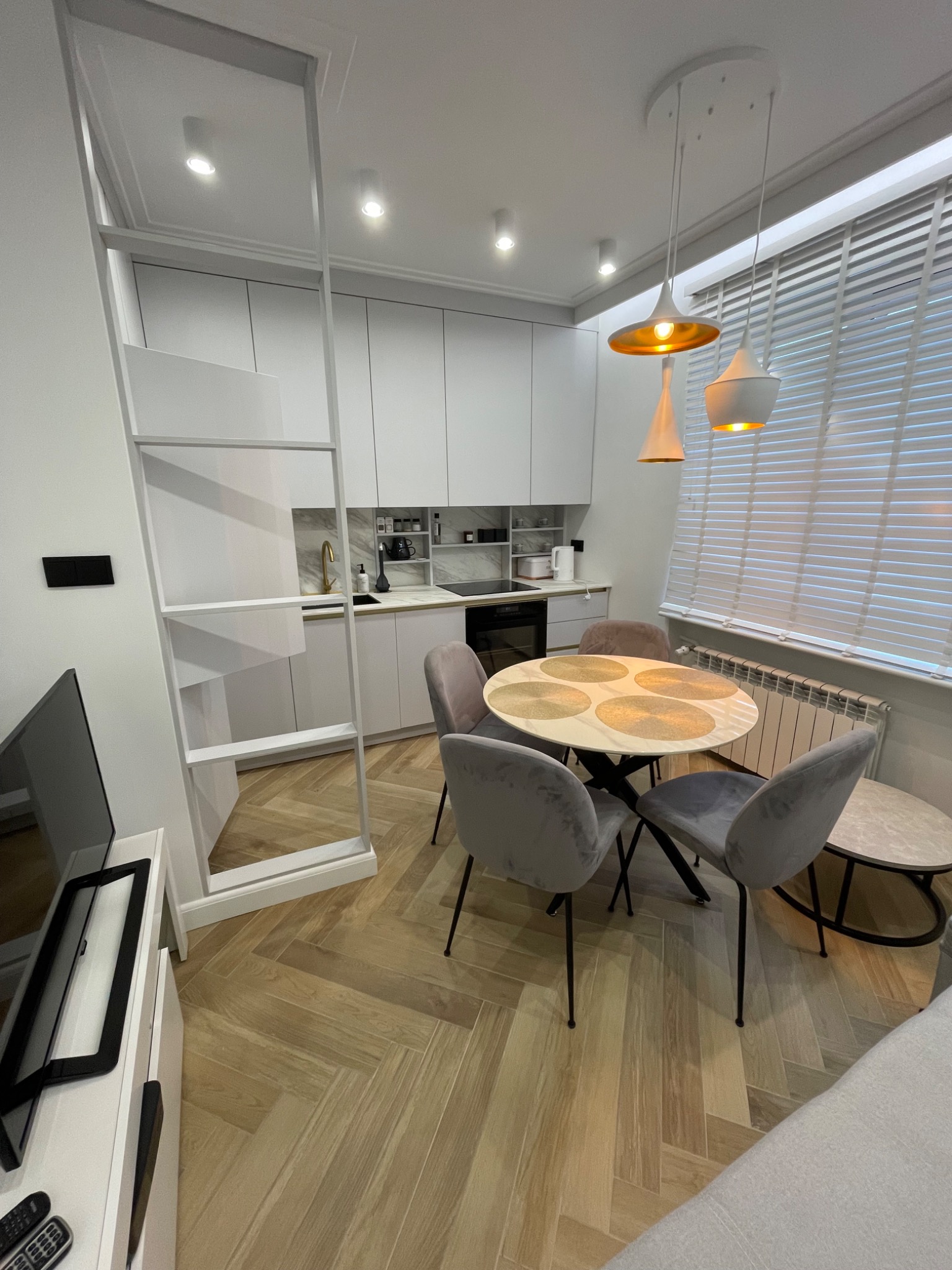 Nowoczesne wnętrze mieszkania z aneksem kuchennym, okrągłym stołem i szarymi krzesłami, oświetlone wiszącymi lampami i światłem wpadającym przez żaluzje.