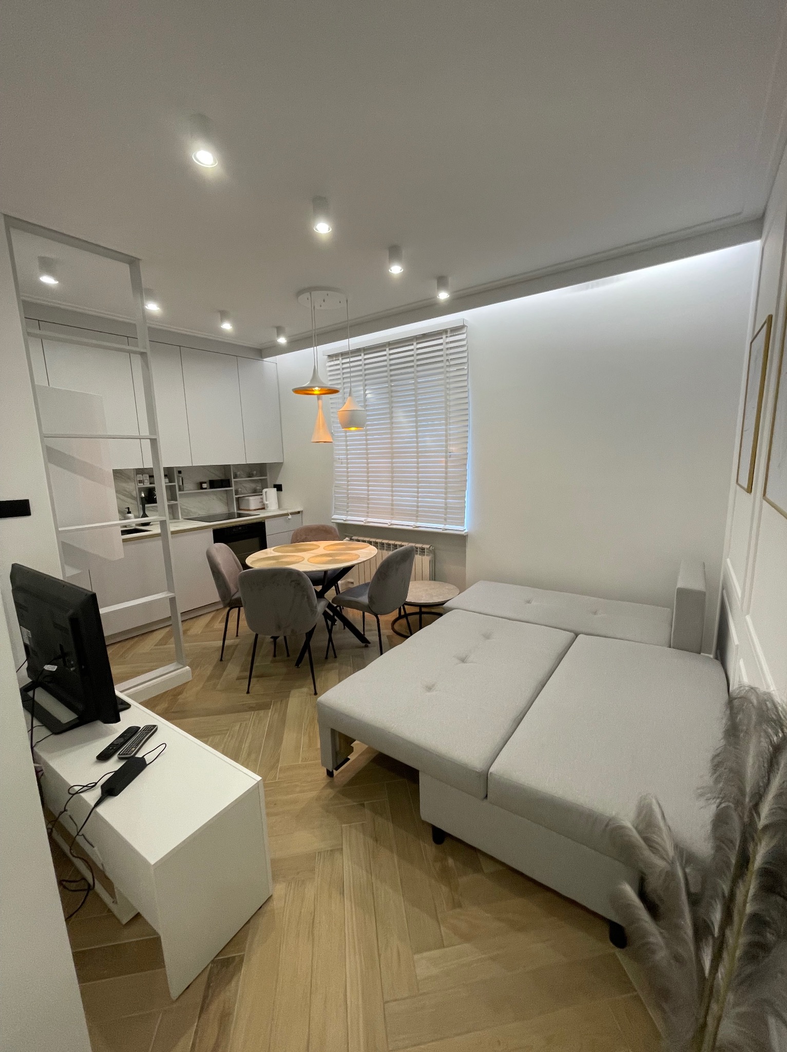 Jasne, nowocześnie urządzone mieszkanie z aneksem kuchennym, okrągłym stołem z krzesłami i rozkładaną sofą narożną na parkiecie w jodełkę.