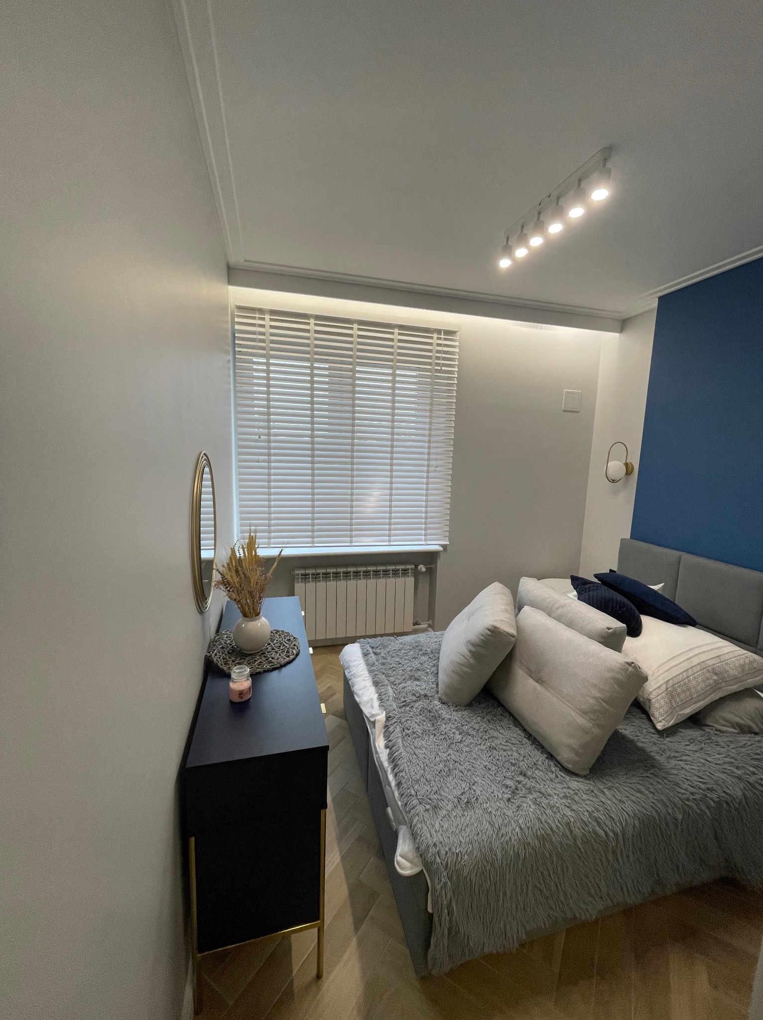 Nowoczesna sypialnia z łóżkiem z szarą narzutą, ciemnoniebieską ścianą akcentującą, czarną komodą z dekoracjami i białymi żaluzjami w oknie.