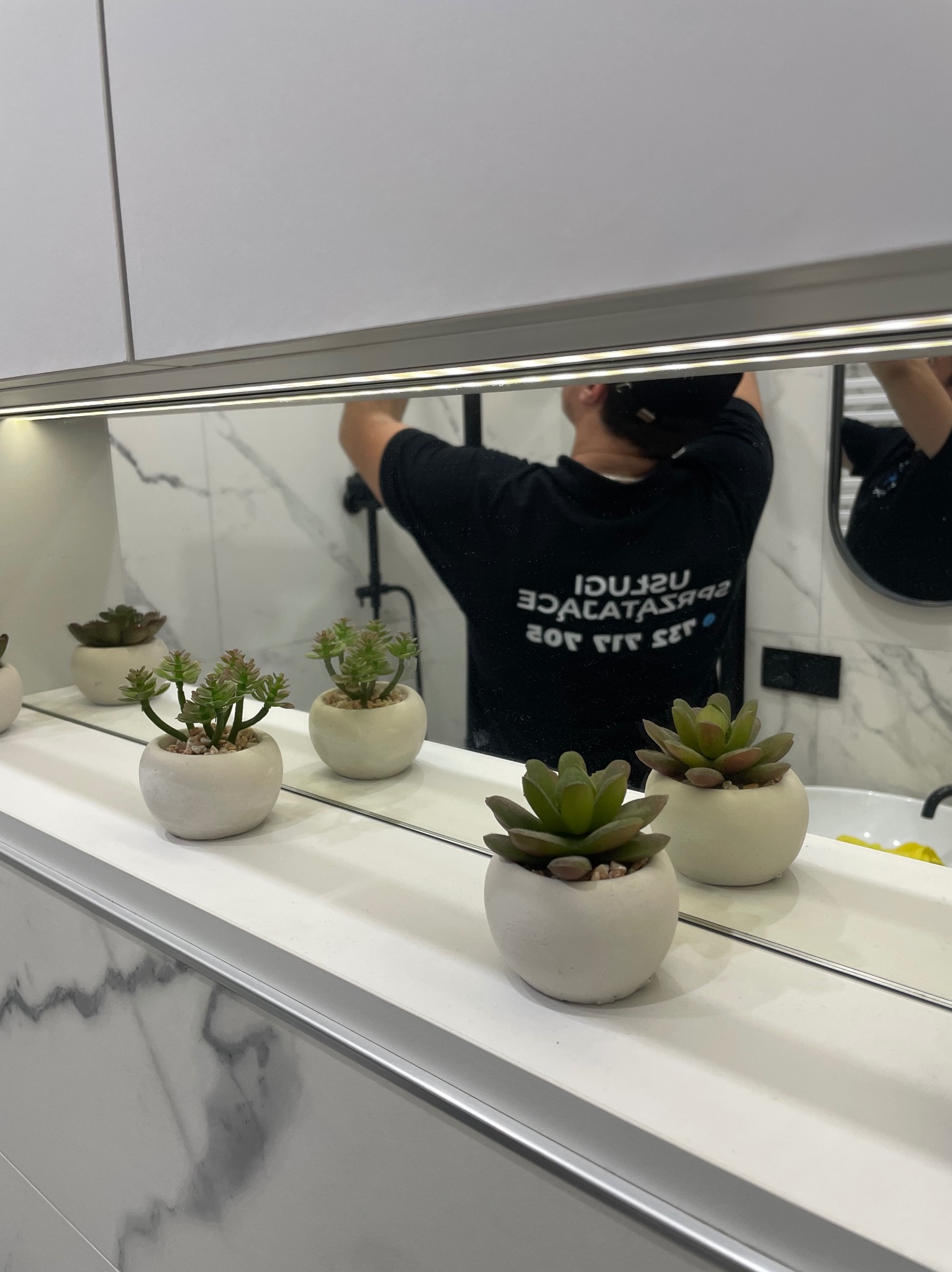 Odbicie w lustrze łazienki: mężczyzna w czarnej koszulce z napisem, myjący lustro nad umywalką, na półce przed lustrem ustawione dekoracyjne sukulenty w białych doniczkach.