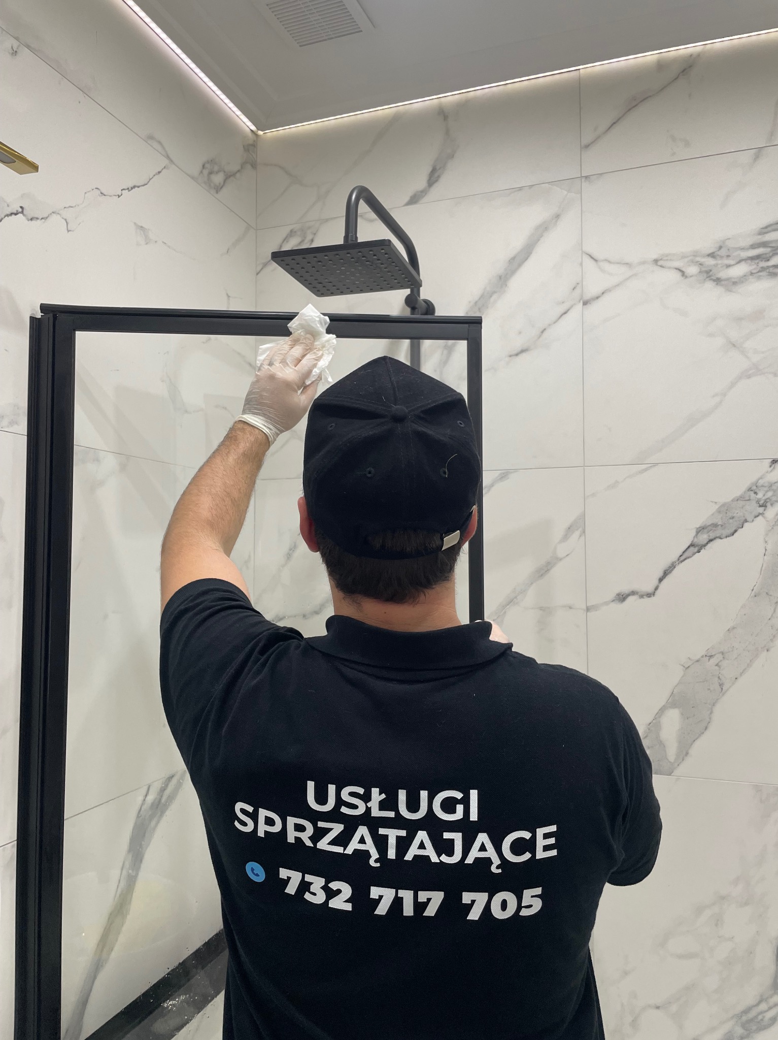 Osoba w czarnej koszulce z napisem 'Usługi Sprzątające' czyści szklaną kabinę prysznicową w łazience z marmurowymi płytkami, używając białej ściereczki i rękawic ochronnych.