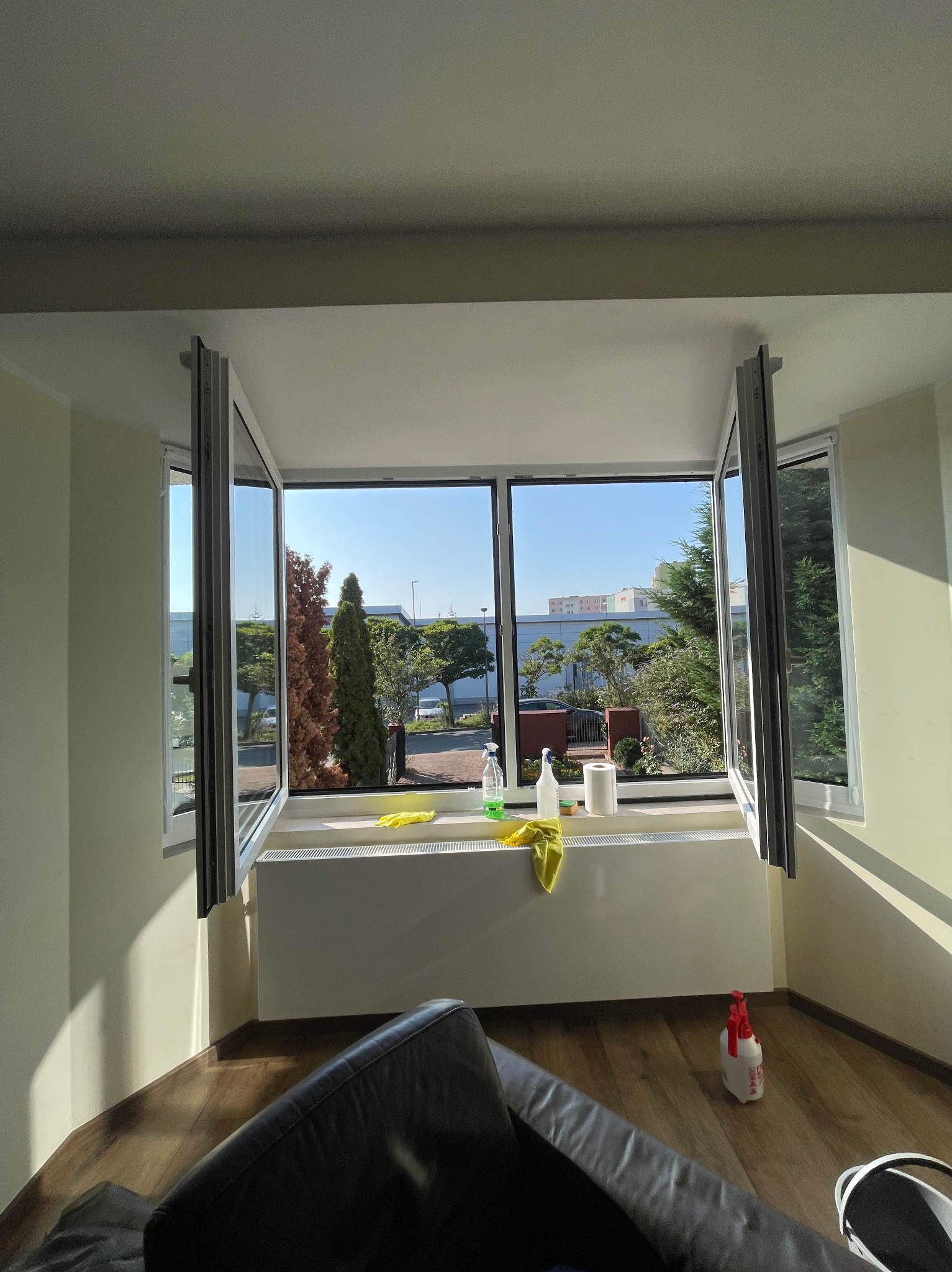 Otwarty okno z widokiem na zieleń w Bydgoszczy, środki czystości i żółte ściereczki na parapecie, butelka ze spryskiwaczem na podłodze.