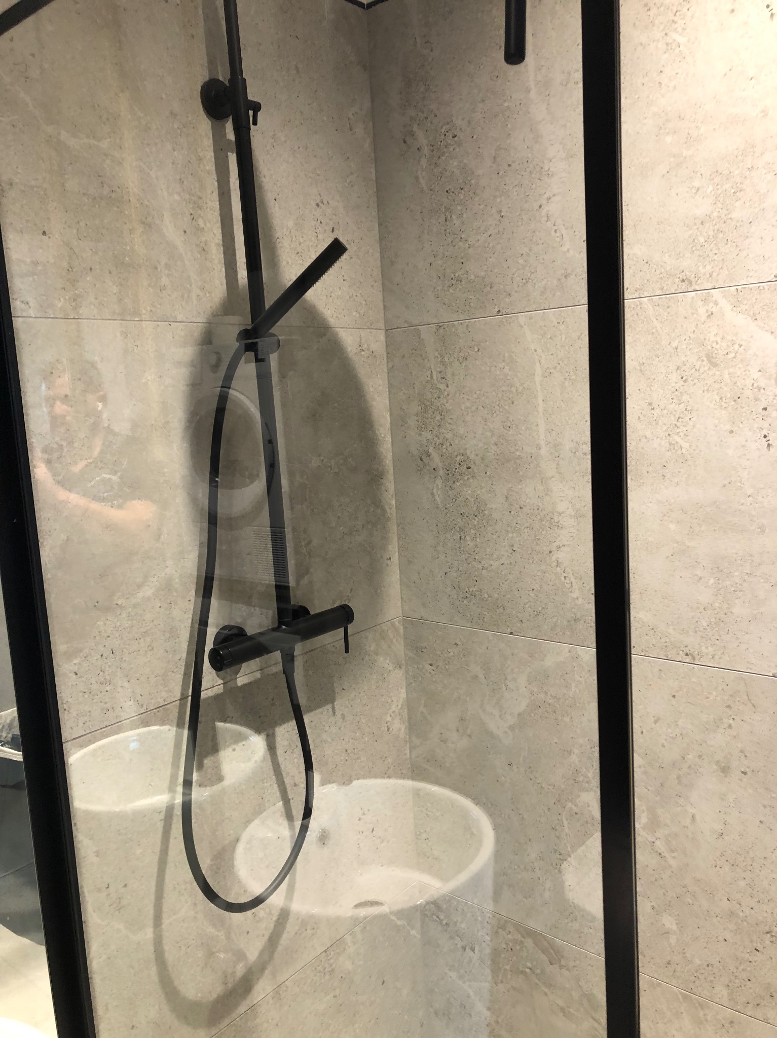 Nowoczesna kabina prysznicowa z czarną armaturą na tle płytek imitujących kamień, widoczna umywalka ceramiczna za szybą.