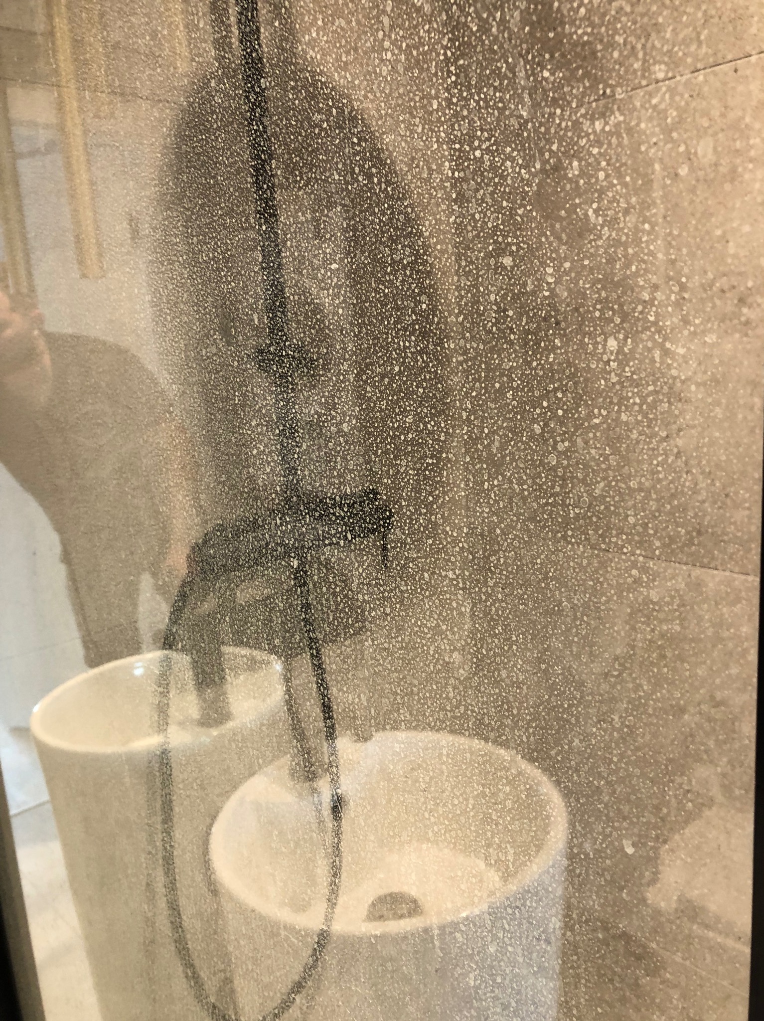 Zabrudzona kabina prysznicowa z widocznymi kroplami wody i osadem, białe umywalki w tle, osoba czyszcząca widoczna jako rozmazana sylwetka po lewej stronie.