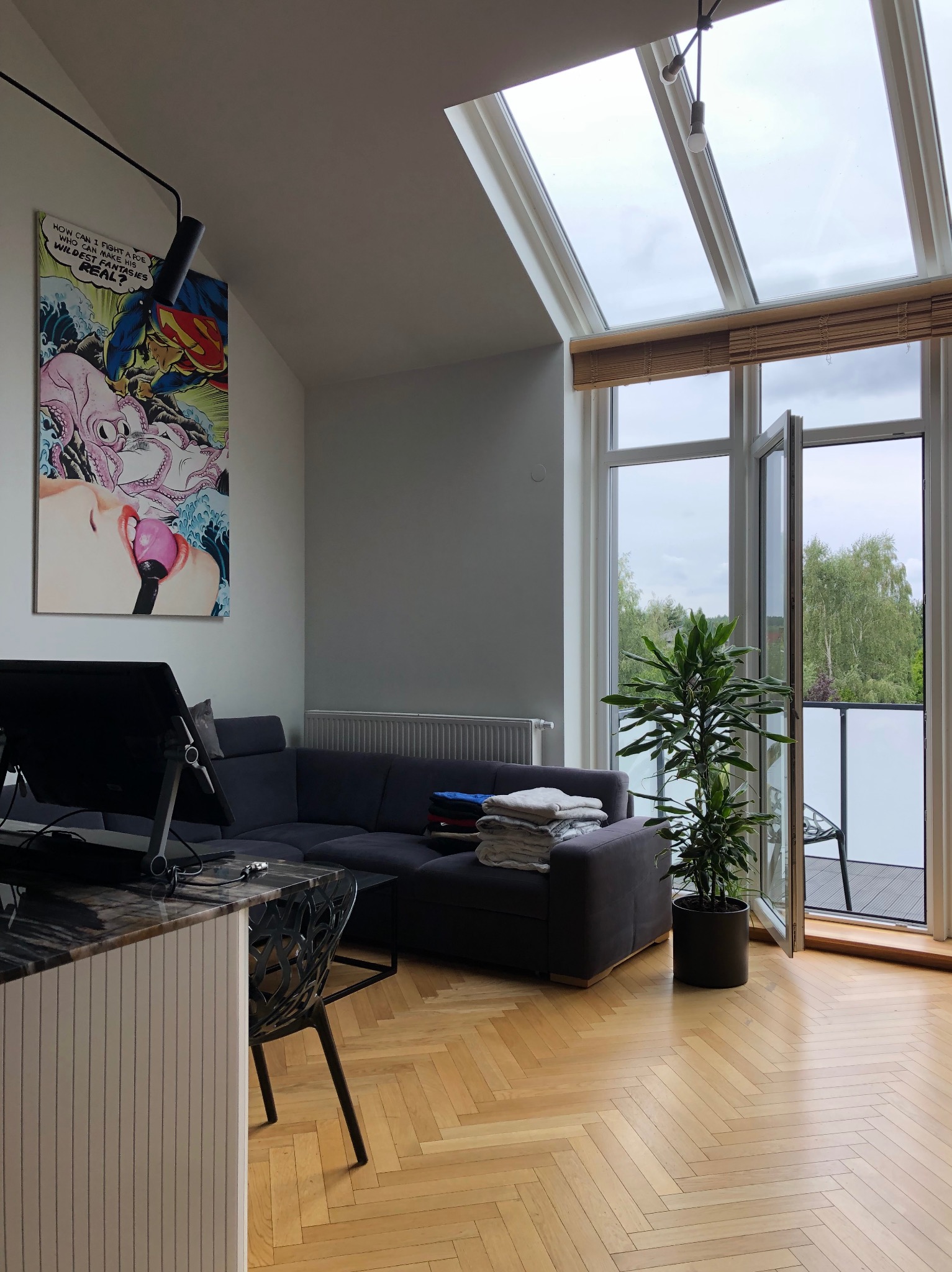 Jasne wnętrze mieszkania z parkietem w jodełkę, szarą sofą z ułożonymi ręcznikami, dużym oknem balkonowym z widokiem na zieleń i komiksem na ścianie.