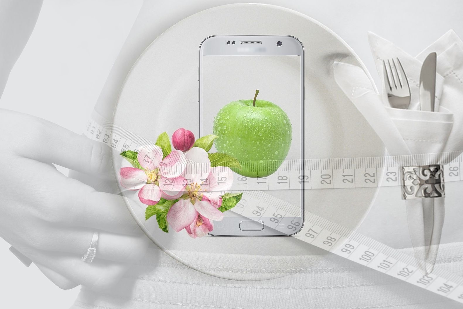 Kompozycja z zielonym jabłkiem na ekranie smartfona, kwiatami jabłoni i miarką krawiecką na białym talerzu, symbolizująca zdrowe odżywianie i dbanie o sylwetkę.