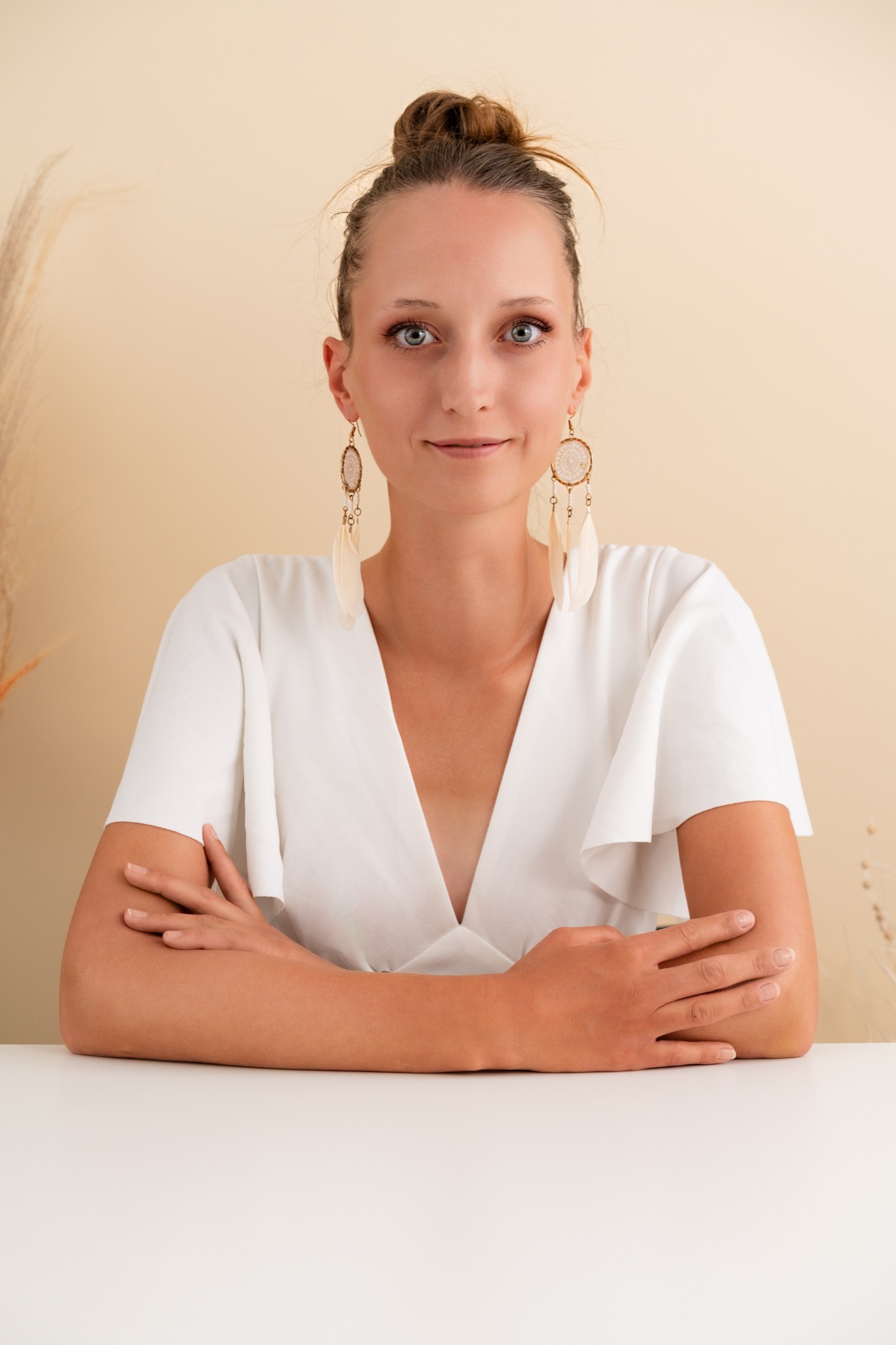 Portret kobiety o jasnych oczach i włosach upiętych w kok, z kolczykami w stylu boho, ubrana w białą bluzkę z dekoltem V, siedzącej za białym stołem.
