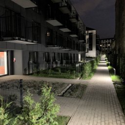 Klaudia Grzesik L-KG Instalacje Elektryczne - Oświetlona ścieżka prowadząca wzdłuż nowoczesnego budynku mieszkalnego nocą. Lampy oświetlają chodnik, tworząc przyjemną atmosferę.